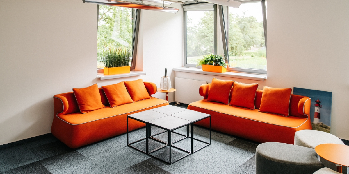 Kreativ-Lounge Seeblick. Ein orangefarbenes Sofa, orangefarbene Blumentöpfe und ein Blick aus dem Fenster auf den Eventgarten.