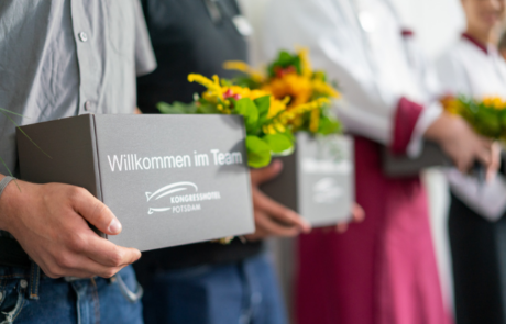 Jemand hält eine Geschenkbox mit der Aufschrift "Willkommen im Team" und einen Blumenstrauß in der Hand.